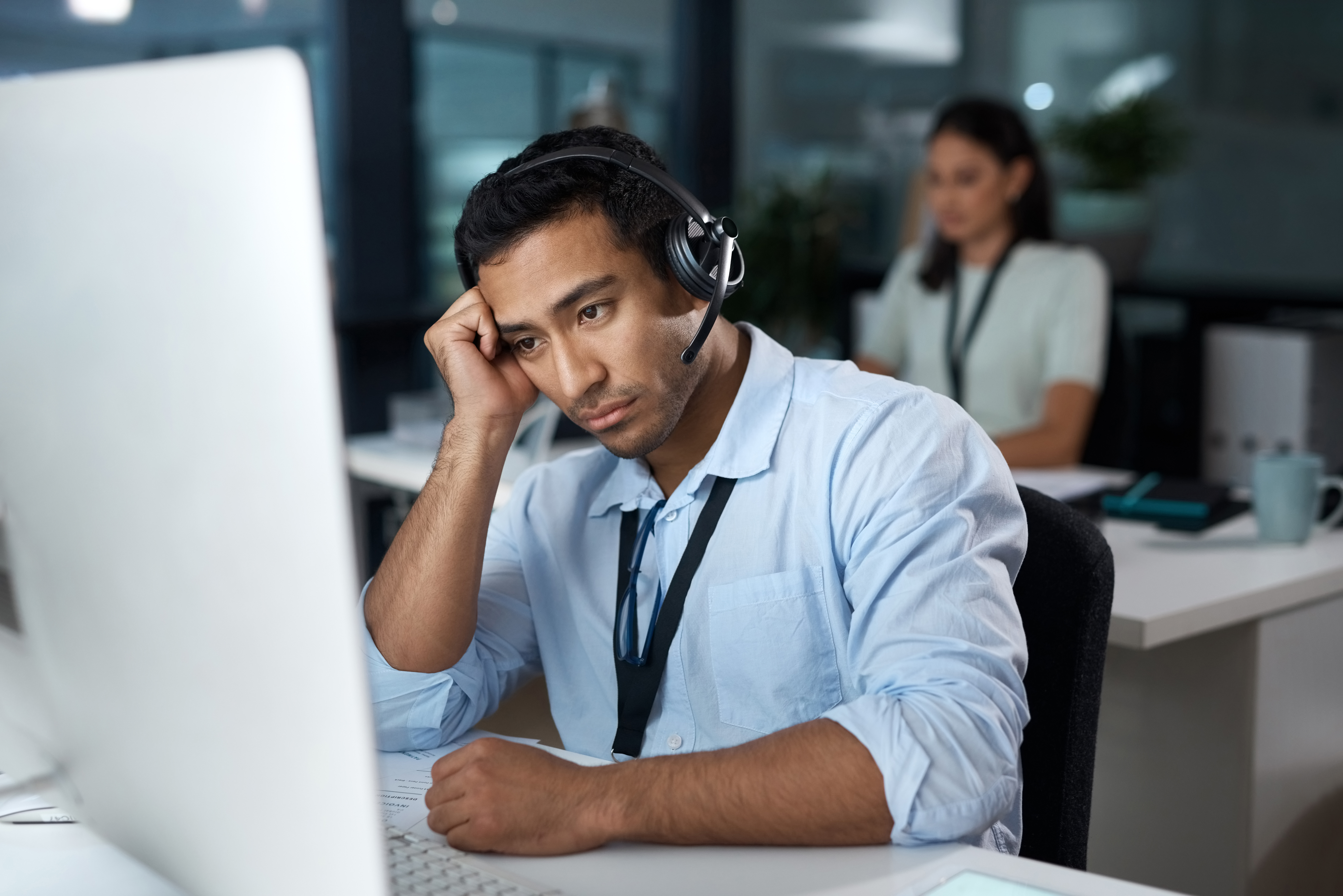 Man at desk at work wearing a headset and staring forlorn at his computer